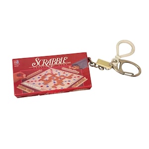 May include: A miniature Scrabble crossword game box keychain featuring a red rectangular package with the Scrabble logo and a game board illustration. The keychain includes a silver metal ring and a white plastic clip attached by a short chain.