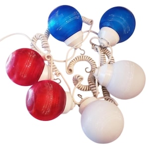 May include: A string of decorative lights with red, white, and blue globe-shaped bulbs. The bulbs are attached to a white, swirled wire frame. The lights are reminiscent of patriotic holiday decorations.