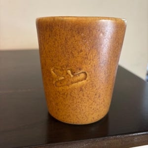 Puede incluir: Una taza cilíndrica de cerámica con un esmalte moteado en tonos marrón y bronceado. La superficie exterior presenta una pequeña forma abstracta grabada que se asemeja a un motivo de ave o ala estilizada en el centro.