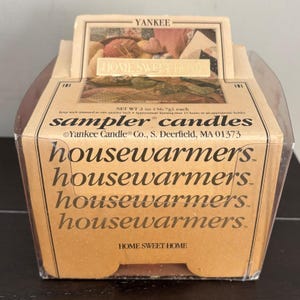 May include: A Yankee Candle sampler candle in a clear plastic container with a tan cardboard label. The label features the text Home Sweet Home, sampler candles, and housewarmers. It notes a net weight of 56.7g and a 15-hour burn time.