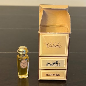 Puede incluir: Un frasco de perfume de vidrio en miniatura con tapón dorado y una etiqueta que dice Calèche Hermès Paris, acompañado de su caja de cartón rectangular original con el nombre de la marca Hermès y un gráfico de un carruaje.