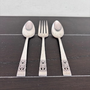 Puede incluir: Un juego de tres utensilios de color plateado: dos cucharas y un tenedor. Las cucharas y el tenedor tienen un diseño floral decorativo en los mangos. Los utensilios están colocados sobre una superficie de madera oscura.