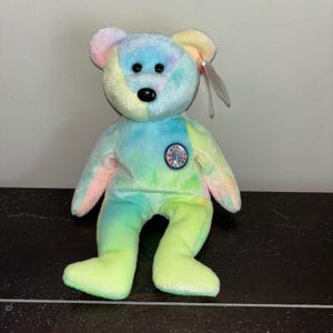 Può includere: Un colorato orsacchiotto di peluche con un motivo tie-dye in tonalità pastello di rosa, giallo, blu e verde. L'orso ha occhi a bottone neri, un naso nero e una piccola toppa circolare sul petto. Un'etichetta è attaccata all'orecchio.