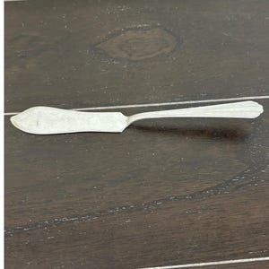 May include: A silver butter knife with a wide, flat blade and a long, slender handle. The handle has a decorative, ridged design. The knife is made of a shiny, metallic material and is designed for spreading butter or other soft spreads.