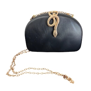 May include: Black clutch with a gold snake embellishment and a gold chain strap. The clutch has a textured surface and a curved shape. The snake is detailed with scales and a head. The chain is a delicate link style.