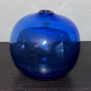May include: A round, cobalt blue glass float with a small opening at the top. The surface of the float is smooth and reflective, with highlights and shadows. The object is sitting on a dark surface.