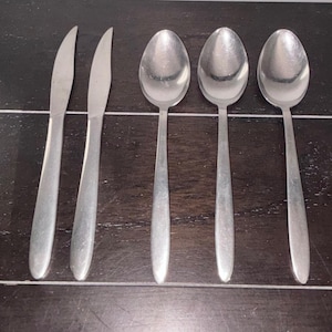 May include: A collection of stainless steel cutlery, comprising two knives and three spoons. The utensils are displayed on a dark wooden surface. The knives have a pointed blade, and the spoons have an oval bowl.