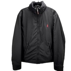 May include: Black zip-up jacket with a high collar and a red logo on the chest. The jacket has two front pockets and a full-length zipper. The jacket is made of a smooth, water-resistant material.