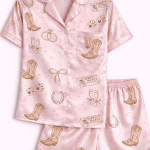 May include: Pale pink pyjama set featuring a button-down shirt and shorts. The set is patterned with cowboy boots, dice, horseshoes, champagne flutes, and the text "PERFECT MATCH".