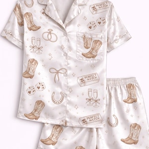 May include: Cream satin pyjama set featuring a repeating pattern of cowboy boots, dice, champagne glasses, horseshoes, and the text "Perfect Match". The set includes a button-down shirt and shorts.