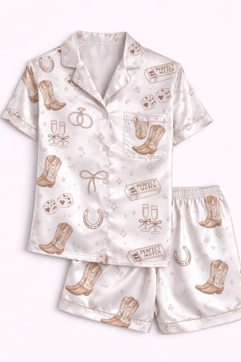 May include: Cream satin pyjama set featuring a repeating pattern of cowboy boots, dice, champagne glasses, horseshoes, and the text "Perfect Match". The set includes a button-down shirt and shorts.