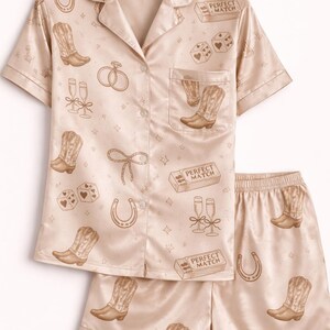 May include: A light beige pajama set with a button-down shirt and shorts. The set is patterned with cowboy boots, horseshoes, dice, champagne flutes, and the text "PERFECT MATCH".