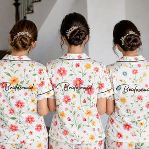 May include: Three people wearing matching floral print pajama sets. The white sets feature a colorful flower pattern with pink, yellow, and blue blooms. The word "Bridesmaid" is printed on the back of each shirt.