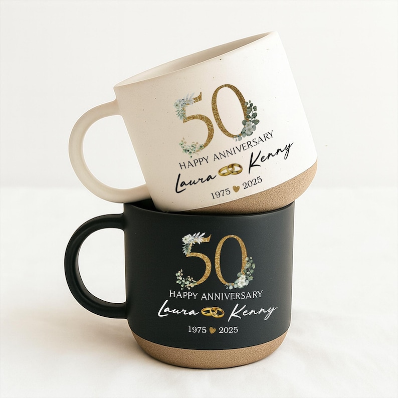 May include: Two stacked ceramic mugs. The top mug is white with a tan base, the bottom mug is black with a tan base. Both mugs have a gold "50" with floral accents, the words "Happy Anniversary", names, and the years "1975 2025".