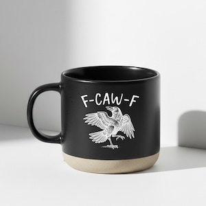 May include: Black ceramic mug with a white illustration of a raven with outstretched wings and the text "F-CAW-F". The mug has a beige base and a black handle. The design is a humorous take on a bird.