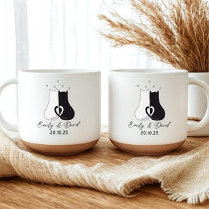 Può includere: Due tazze in ceramica bianca con base marrone. Ogni tazza presenta un disegno di gatto bianco e nero con cuori e i nomi "Emily & David" e la data "20.10.25". Le tazze sono su un tessuto beige.