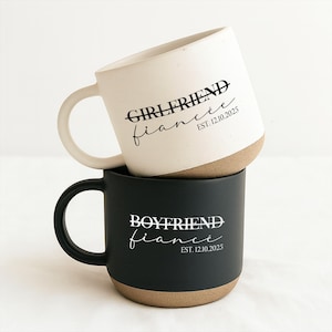 Puede incluir: Dos tazas de cerámica apiladas. La taza superior es blanca con el texto "GIRLFRIEND fiancé EST. 12.10.2025" en negro. La taza inferior es negra con el texto "BOYFRIEND fiancé EST. 12.10.2025" en blanco. Ambas tazas tienen una base marrón.