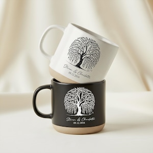 May include: Two ceramic mugs, one white and one black, stacked. Each mug features a black tree design with the names "Steven & Charlotte" and the date "09.12.2016" printed below the tree. The base of each mug is a tan colour.