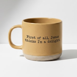 May include: A mustard yellow ceramic mug with a speckled finish and a light gray unglazed base. The front features black typewriter-style text that reads: First of all, Jesus thinks I'm a delight. The mug has a curved handle on the left side.