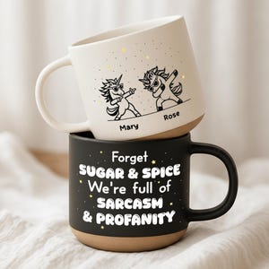 Puede incluir: Dos tazas de cerámica apiladas. La taza superior es blanca con un diseño de unicornio negro y los nombres "Mary" y "Rose". La taza inferior es negra con el texto "Forget Sugar & Spice We're full of Sarcasm & Profanity".