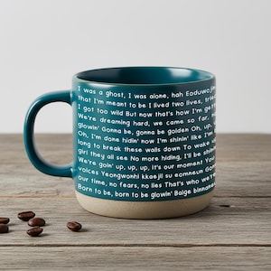 May include: Teal ceramic mug with a beige base, featuring white text. The mug has a large handle and is filled with a poem. Coffee beans are scattered on the wooden surface.
