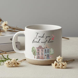 May include: White ceramic mug with a tan base and handle. The mug features a watercolor illustration of two houses, trees, and the text "I wish you lived next door" in black script. Pink hearts are also included.