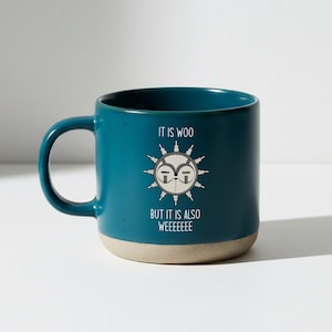 May include: Teal ceramic mug with a beige base and a large handle. The mug features a graphic of a sun-like design with glasses and the text "IT IS WOO BUT IT IS ALSO WEEEEEEE".