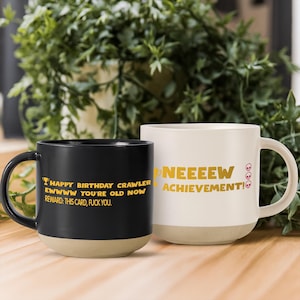 May include: Two ceramic mugs with humorous text. One is black with gold text that reads "Happy Birthday Crawler Ewwww You're Old Now Reward: This Card, Fuck You." The other is white with gold text that says "Neeeeew Achievement!" and skull emojis.