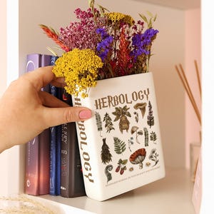 Könnte beinhalten: Eine weiße Keramikvase in Buchform mit dem Wort "HERBOLOGY" und botanischen Illustrationen. Die Vase ist mit bunten Trockenblumen gefüllt, darunter Gelb, Lila und Rot.