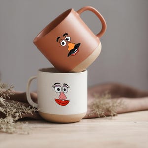 Può includere: Due tazze in ceramica, una impilata sull'altra. La tazza superiore è marrone con la faccia di un uomo dei cartoni animati, e la tazza inferiore è bianca con la faccia di una donna dei cartoni animati. Entrambe le tazze hanno una base beige e un manico.