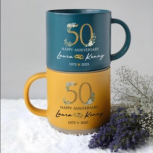 May include: Two ceramic mugs, one teal and one yellow, stacked on top of each other. Each mug features a gold "50" with floral accents, the words "Happy Anniversary", names, and the years "1975 2025".