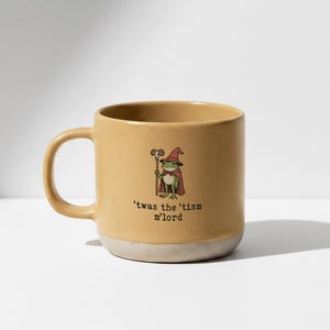 May include: Mustard yellow ceramic mug with a white base. The mug features a cartoon frog dressed as a wizard, holding a staff, and wearing a red hat and cape. The text on the mug reads, "'twas the 'tism m'lord."