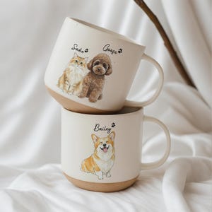 Op de afbeelding: Twee gestapelde keramische mokken met illustraties van huisdieren. De bovenste mok heeft een kat en een hond met de namen "Sadie" en "Goose". De onderste mok heeft een corgi-illustratie met de naam "Bailey". Elke mok heeft een beige basis en een witte bovenkant.