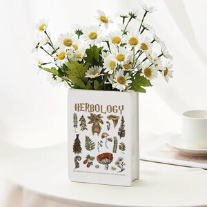 May include: A white rectangular vase with the word "HERBOLOGY" in gold lettering, featuring botanical illustrations. The vase holds a bouquet of white daisies with yellow centers. The text "HOGWARTS SCHOOL OF WITCHCRAFT AND WIZARDRY" is at the bottom.