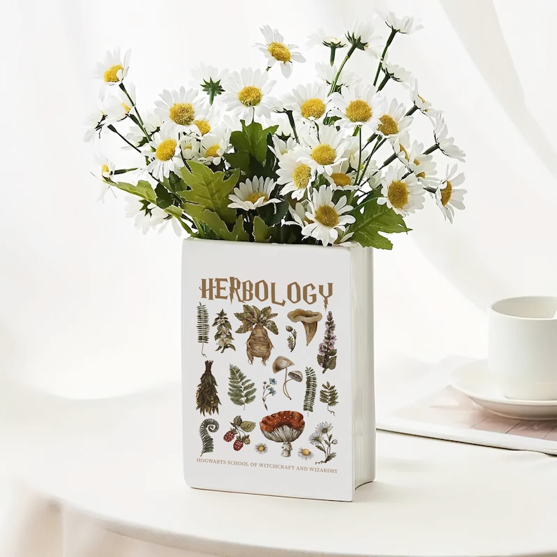 May include: A white rectangular vase with the word "HERBOLOGY" in gold lettering, featuring botanical illustrations. The vase holds a bouquet of white daisies with yellow centers. The text "HOGWARTS SCHOOL OF WITCHCRAFT AND WIZARDRY" is at the bottom.