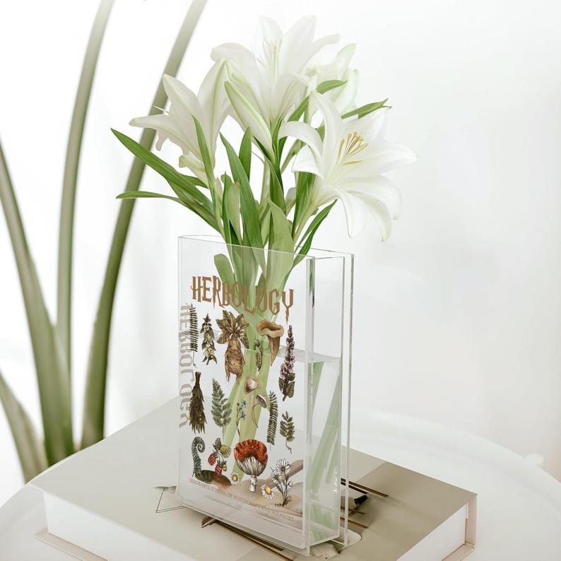 May include: A clear acrylic vase with the word "HERBOLOGY" in gold lettering, filled with white lilies. The vase has botanical illustrations. The vase is sitting on a white book.