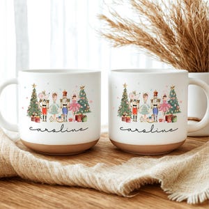Może przedstawiać: Dwa białe ceramiczne kubki z brązową podstawą. Każdy kubek przedstawia akwarelową ilustrację sceny bożonarodzeniowej z myszą, dziadkiem do orzechów i choinką. Imię "Caroline" jest napisane poniżej.