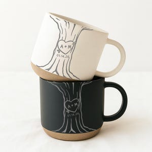 May include: Two ceramic mugs, one cream and one black, stacked. Each mug has a tree design with a heart and arrow, the initials "A + M", and the date "01.14.26". The mugs have a natural, unglazed base and a curved handle.