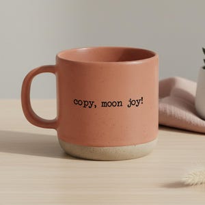 Peut inclure: Une tasse en céramique de couleur terracotta tachetée, avec une base beige et une anse incurvée. Le texte "copy, moon joy!" est imprimé en noir. La tasse est posée sur une surface en bois clair.