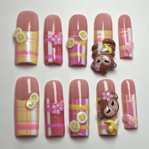 Könnte beinhalten: Ein Set aus zehn künstlichen Nägeln mit einem rosa-gelben Karomuster. Einige Nägel weisen 3D-Verzierungen von Blumen, Bananen und einem Affengesicht auf. Die Nägel haben eine abgerundete Form und eine rosa Basis.