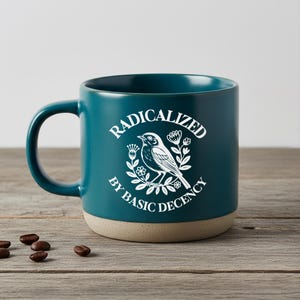 Può includere: Tazza in ceramica color verde acqua con grafica bianca di uccello e fiori, e la scritta "RADICALIZED BY BASIC DECENCY". La tazza ha una base beige e un manico curvo. Chicchi di caffè sparsi sulla superficie di legno.