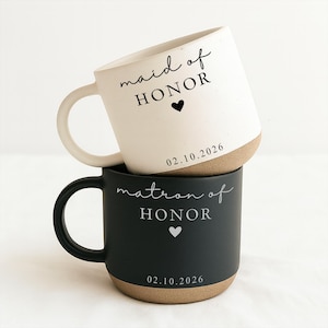 Puede incluir: Dos tazas de cerámica apiladas. La taza superior es blanca con las palabras "maid of HONOR", un corazón y la fecha "02.10.2026" en negro. La taza inferior es negra con "matron of HONOR", un corazón y la fecha "02.10.2026" en blanco.