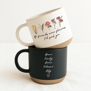 May include: Two ceramic mugs stacked on top of each other. The top mug is white with floral illustrations and the text "If friends were flowers, I'd pick you." The bottom mug is black with names and a heart.