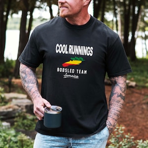 May include: Black t-shirt with the words "COOL RUNNINGS" above a graphic of a bobsled and the text "BOBSLED TEAM Jamaica". The shirt is short-sleeved and has a crew neck. A person is holding a dark blue travel mug.