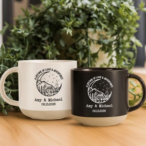 Pode incluir: Duas canecas de cerâmica, uma creme e outra preta, com um design temático de acampamento. Cada caneca apresenta o texto "A Lifetime of Love & Adventures", os nomes "Amy & Michael" e a data "09.15.2026".