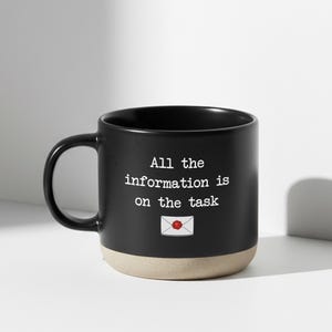 Może przedstawiać: Czarny ceramiczny kubek z beżową podstawą i czarnym uchwytem. Kubek zawiera biały napis "All the information is on the task" oraz małą grafikę zapieczętowanej koperty.