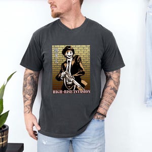 Puede incluir: Camiseta gris oscuro con un gráfico de "High-Rise Invasion". El diseño presenta un personaje con traje y sombrero que sostiene un arma, sobre un fondo de pared de ladrillos. El texto "HIGH-RISE INVASION" está debajo de la imagen.