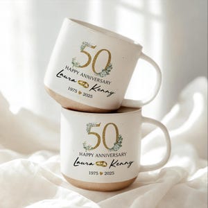May include: Two white ceramic mugs stacked on top of each other. Each mug has a gold "50" with floral accents, the words "Happy Anniversary", and the names "Laura & Kenny" with the years "1975 2025". The bottom of the mugs are tan.