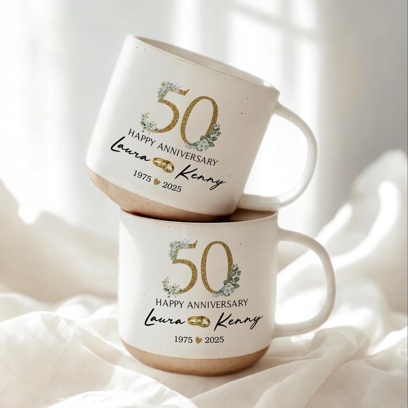 May include: Two white ceramic mugs stacked on top of each other. Each mug has a gold "50" with floral accents, the words "Happy Anniversary", and the names "Laura & Kenny" with the years "1975 2025". The bottom of the mugs are tan.