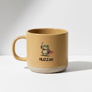 Op de afbeelding: Een beige keramische koffiemok met een cartoon-illustratie van een kikker met een cape en zwaard. Het woord "Huzzah" staat eronder. De mok heeft een beige basis en een gebogen handvat.